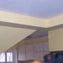 Wnętrze w trakcie prac wykończeniowych w Poznaniu: żółte ściany z szarym pasem dekoracyjnym pod sufitem, widoczne szafki kuchenne i fragment osoby pracującej.